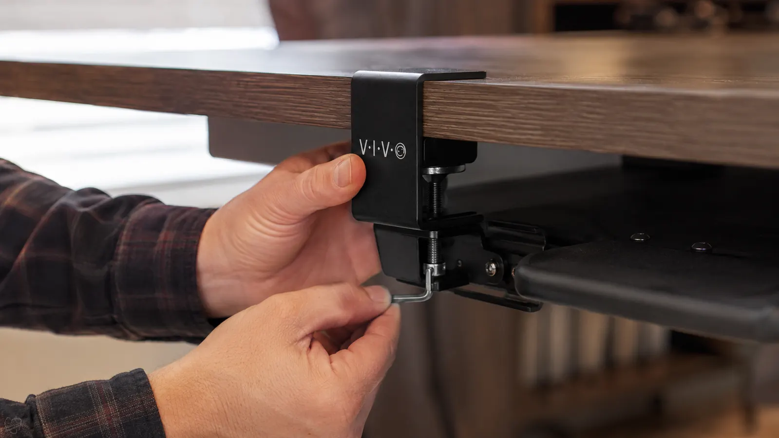 A man's hands clamping a VIVO keyboard tray bracket to the underside of a desk with a hex key