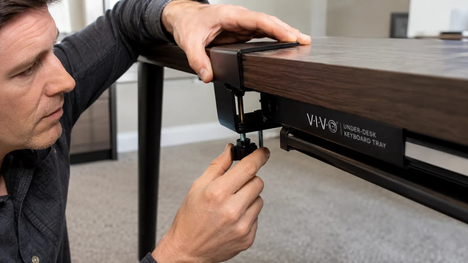 Hand tightening a C-clamp bracket onto the underside of a desk edge to mount a keyboard tray track