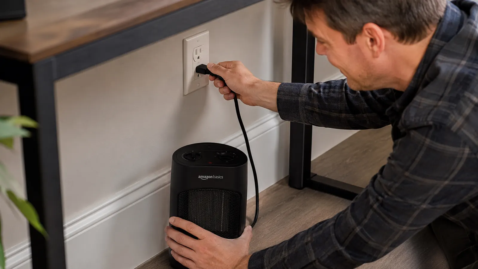 Hand plugging a black Amazon Basics mini space heater directly into a wall outlet under a desk