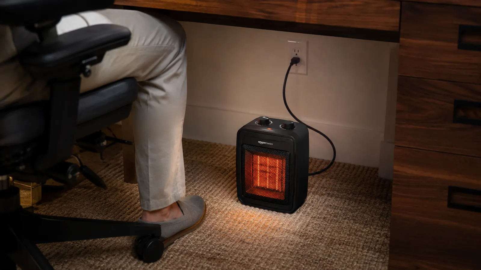 Small black ceramic mini heater plugged in and glowing under a desk, positioned near a person's feet