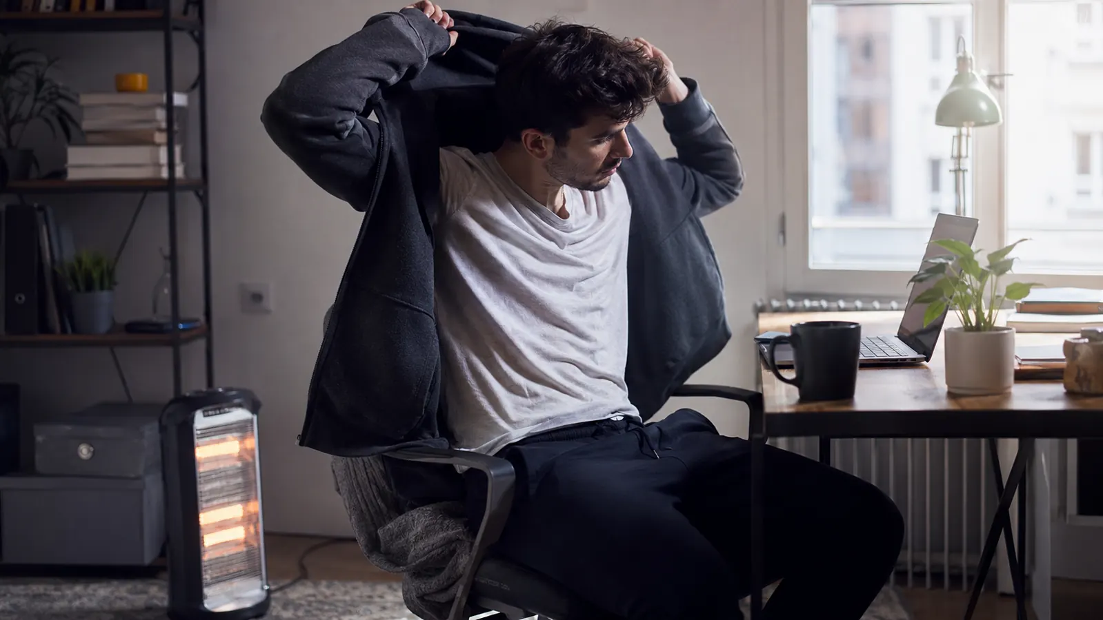 Man in a hoodie removing it while sitting at his desk, small heater visible near his feet