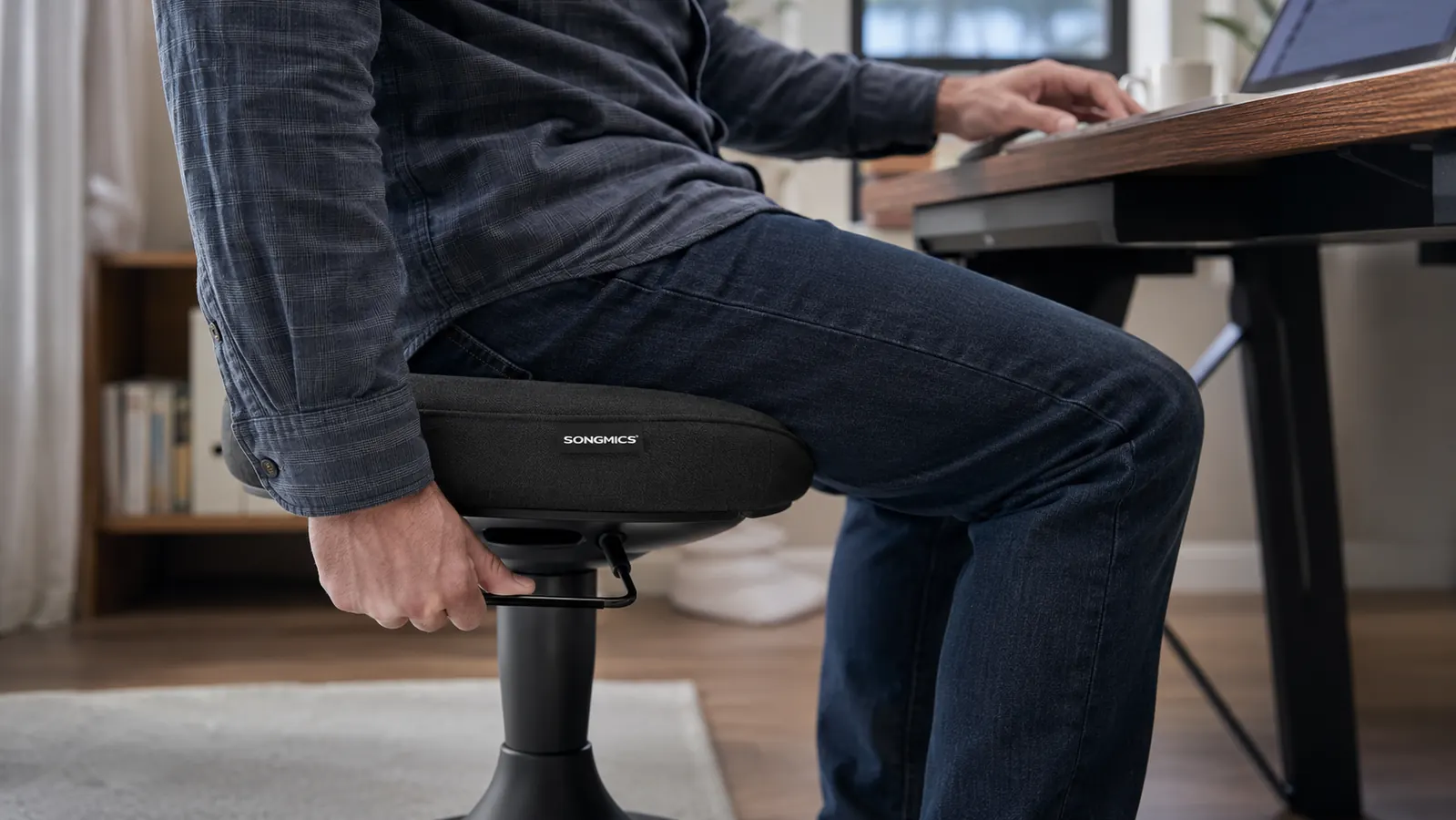 Man adjusting the height lever on a black SONGMICS wobble stool next to his desk